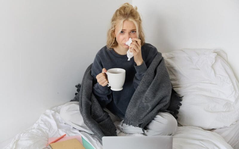 A young woman is at home feeling unwell, sipping tea and passing time in bed