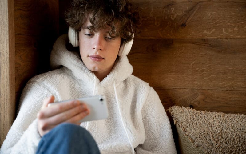 Gen Z teenager sitting on his bed, listening to music on his smartphone