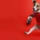 A dog in football gear kicking a field goal like a human against a solid red background. AI-generated image.