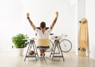 A fortunate freelancer sits at his workspace, surrounded by his hobby items—a longboard, a vintage bicycle, and a green plant—stretching his hand in the air while taking a break.