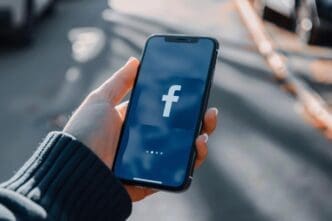 A hand gripping a cell phone displaying the Facebook logo on its screen