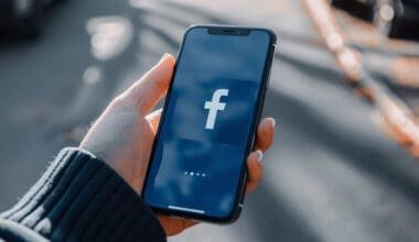 A hand gripping a cell phone displaying the Facebook logo on its screen