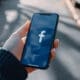 A hand gripping a cell phone displaying the Facebook logo on its screen