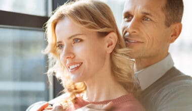 A joyful, upbeat man stands behind his girlfriend, smiling as he embraces her warmly