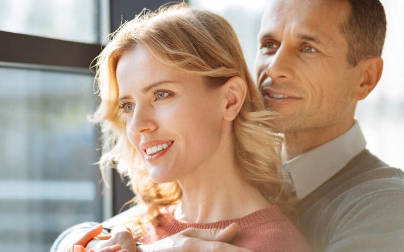 A joyful, upbeat man stands behind his girlfriend, smiling as he embraces her warmly