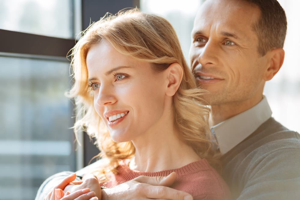A joyful, upbeat man stands behind his girlfriend, smiling as he embraces her warmly