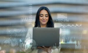 A self-assured young professional woman working on a laptop, with vibrant financial and data job charts superimposed in the background