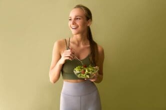 A young athletic woman in a green top and gray leggings is smiling to the side while holding a plate of salad