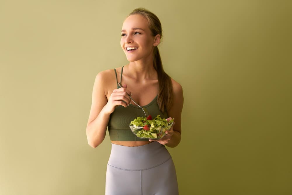 A young athletic woman in a green top and gray leggings is smiling to the side while holding a plate of salad