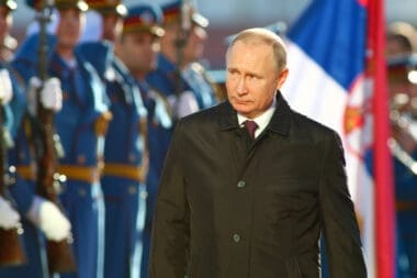 A candid portrait of Vladimir Putin, President of Russia, in a dark coat, with a serious expression, at a formal event in Serbia.