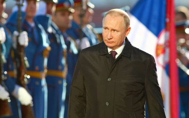 A candid portrait of Vladimir Putin, President of Russia, in a dark coat, with a serious expression, at a formal event in Serbia.