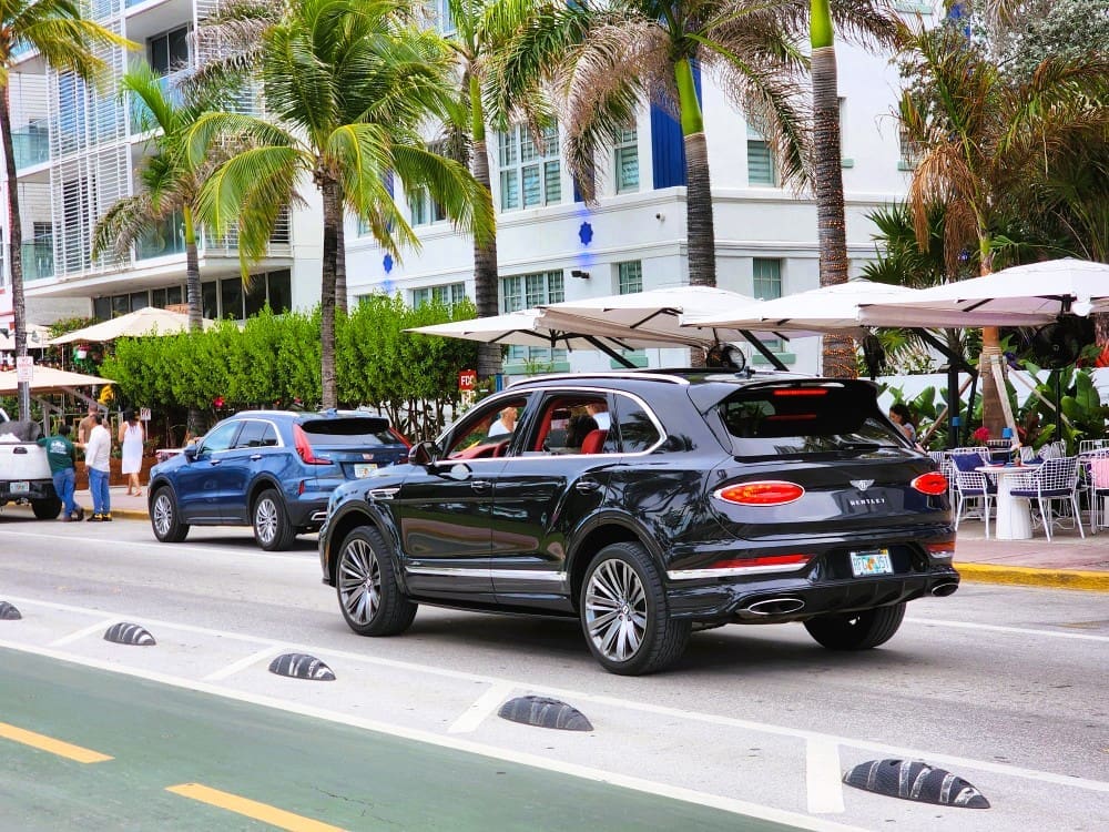 A black Bentley Bentayga SUV drives on Ocean Drive, Miami, passing by outdoor cafes with white umbrellas, palm trees, and art deco buildings on a sunny day.