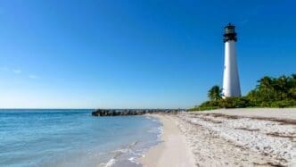 A white lighthouse stands on a sandy beach next to a rock jetty and the ocean under a clear blue sky.