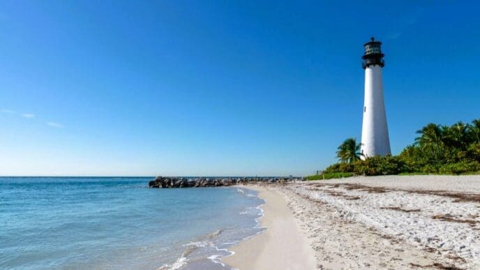 A white lighthouse stands on a sandy beach next to a rock jetty and the ocean under a clear blue sky.