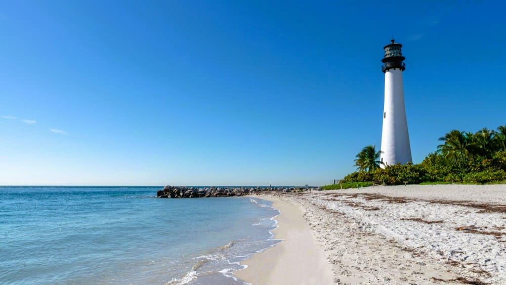 A white lighthouse stands on a sandy beach next to a rock jetty and the ocean under a clear blue sky.