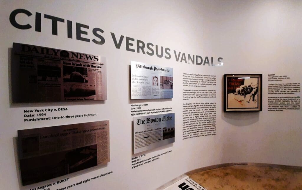 An exhibit wall at the Museum of Graffiti with the large title "CITIES VERSUS VANDALS," featuring various framed newspaper clippings and text detailing legal responses to graffiti, and a framed art piece on the right.