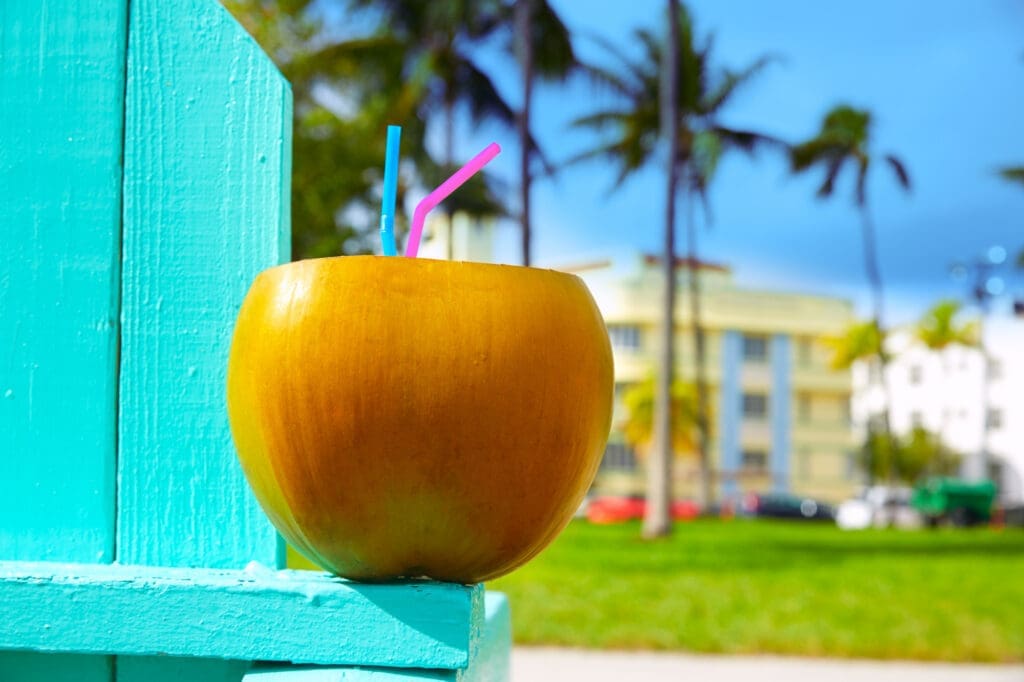 A bright yellow coconut with pink and blue straws sits on a turquoise wooden surface. The background shows a green lawn with palm trees and the blurred Art Deco buildings of Ocean Drive under a blue sky.