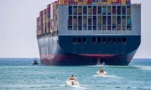 Container ship close to shore, accompanied by a tugboat and smaller vessels