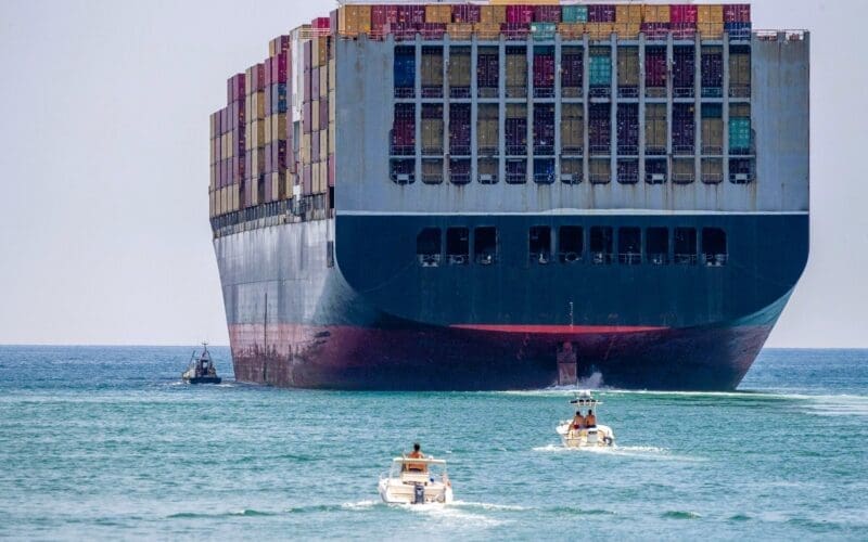 Container ship close to shore, accompanied by a tugboat and smaller vessels