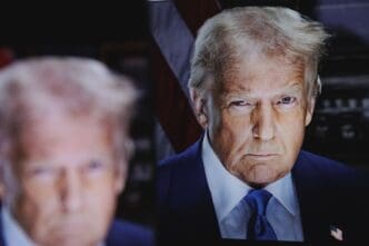 A photograph of a digital screen showing a portrait of President Donald Trump in a dark suit and blue tie, with a serious expression and a blurry reflection of the image in the foreground.