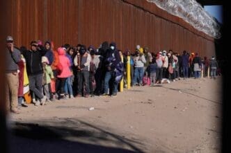 A long line of migrants, including men, women, and children, stands along a tall, rusty border wall with barbed wire on top, waiting in the sun.
