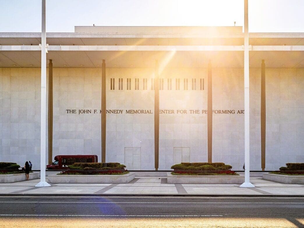 The entrance to the John F. Kennedy Center for the Performing Arts, a white marble building with columns, under a bright sun.