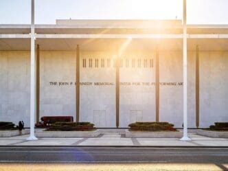 The entrance to the John F. Kennedy Center for the Performing Arts, a white marble building with columns, under a bright sun.