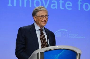 Former Microsoft CEO Bill Gates, smiling and wearing glasses, stands at a podium with a blue background, speaking during a polio eradication funding partnership launch.
