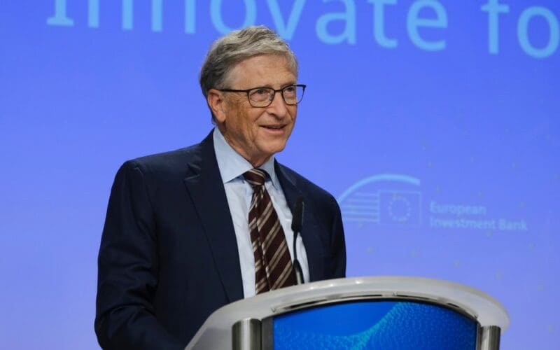 Former Microsoft CEO Bill Gates, smiling and wearing glasses, stands at a podium with a blue background, speaking during a polio eradication funding partnership launch.