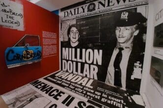 An exhibit at the Museum of Graffiti featuring a blue car door with "CORN BREAD" graffiti, next to a large-scale reproduction of a vintage Daily News headline "MILLION DOLLAR VANDAL" with images of two men, all set against a red wall.
