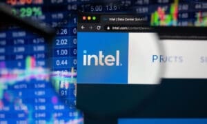 Intel company logo on a website with blurry stock market developments in the background, seen on a computer screen through a magnifying glass.