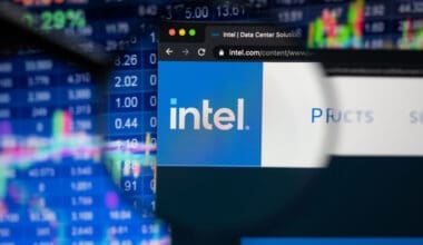 Intel company logo on a website with blurry stock market developments in the background, seen on a computer screen through a magnifying glass.