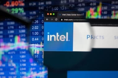 The Intel logo on a website is seen through a magnifying glass, with a blurry stock market chart in the background.