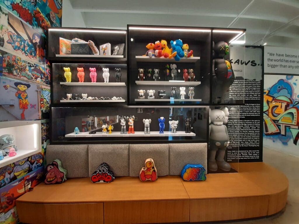 A vibrant display of collectible KAWS art toys and merchandise in various colors and sizes, showcased in illuminated shelves and on a wooden base, surrounded by graffiti art and textual information, within the Museum of Graffiti.