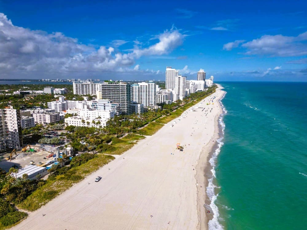A high-angle aerial view of Lummus Park Beach, with a wide stretch of white sand, turquoise ocean waves, a line of tall hotels and residential buildings, and a clear blue sky with white clouds.