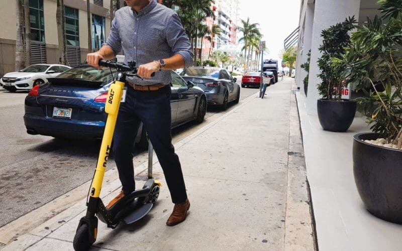 Modern executive man using Bolt, dockless electric scooter, in the streets of Brickell (Miami) with a Porsche behind him