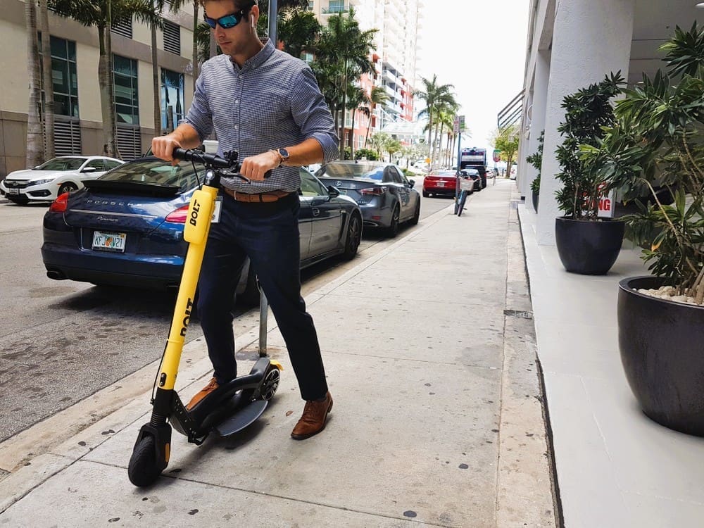 Modern executive man using Bolt, dockless electric scooter, in the streets of Brickell (Miami) with a Porsche behind him