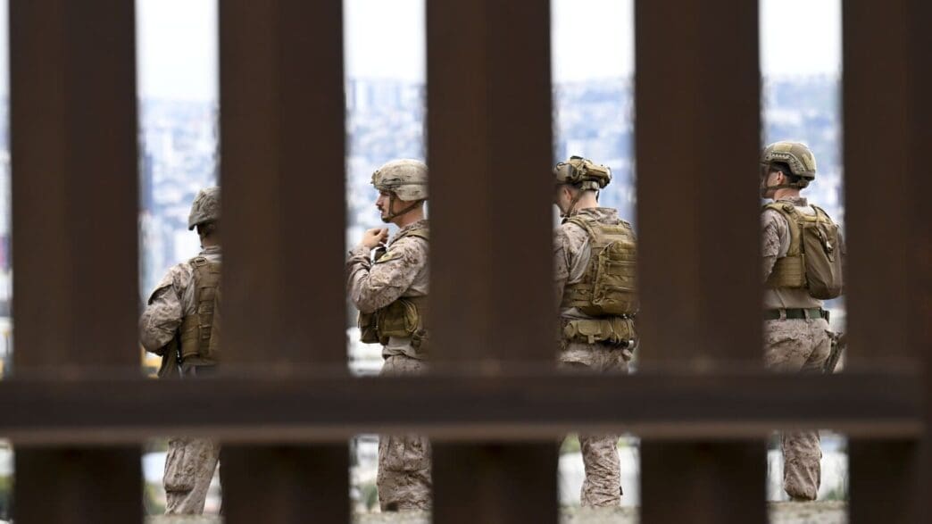 A photograph taken through the vertical slats of a fence, showing four military personnel in full gear standing along the U.S. border wall with a hazy city skyline in the background.