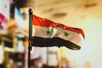 Syria's flag in the foreground with a blurred cityscape background at sunrise