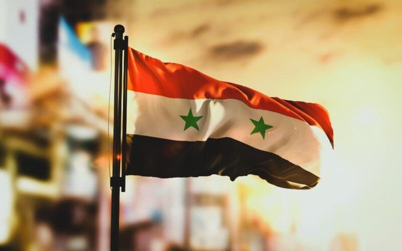 Syria's flag in the foreground with a blurred cityscape background at sunrise