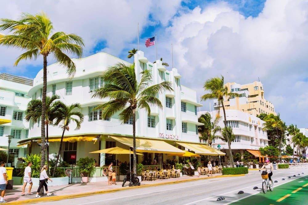 The street view of Ocean Drive in Miami Beach, with a row of iconic white Art Deco hotels with yellow awnings and outdoor seating, a few tall palm trees, and people walking and biking on a sunny day under a blue sky with scattered clouds.