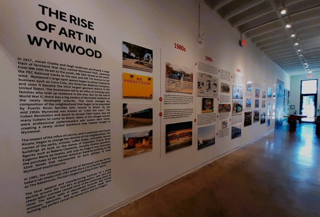 An interior view of the Museum of Graffiti, showing a large wall exhibit titled "THE RISE OF ART IN WYNWOOD" with text and historical photographs illustrating the district's transformation, alongside a timeline for the 1980s, 1990s, and 2000s.