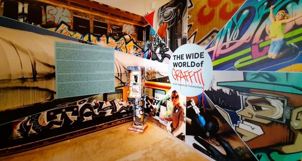 An immersive exhibit at the Museum of Graffiti, featuring a collage of graffiti art, textual information, and a large circular sign that reads "THE WIDE WORLD of GRAFFITI From Vandals to Trendsetters."