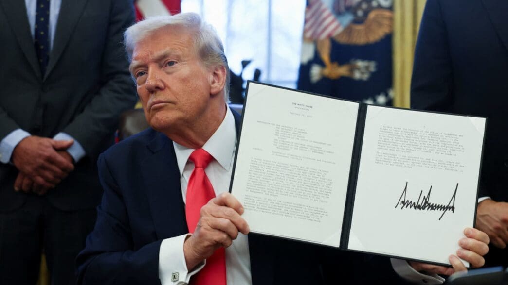 President Donald Trump, wearing a dark suit and red tie, holds up an official document with text visible, likely an executive order or proclamation, during a formal announcement.