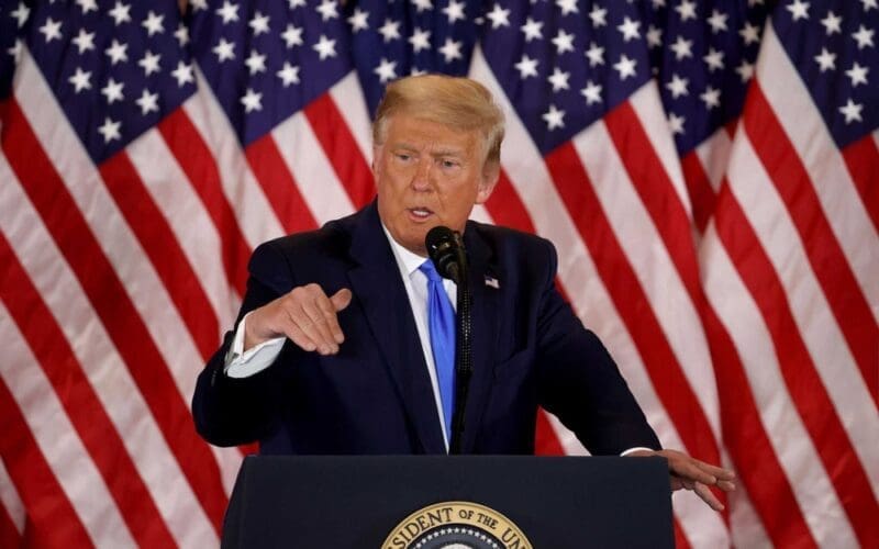President Donald Trump speaking at a podium with multiple American flags in the background.