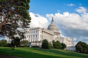 A wide, low-angle view of the majestic U.S. Capitol Building in Washington, D.C., with its iconic dome, white columns, and expansive wings, set against a blue sky with fluffy white clouds and green lawns in the foreground.