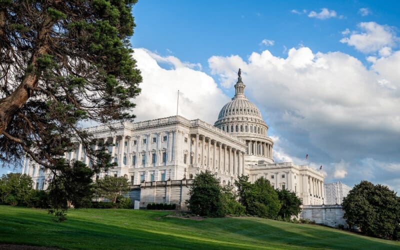 A wide, low-angle view of the majestic U.S. Capitol Building in Washington, D.C., with its iconic dome, white columns, and expansive wings, set against a blue sky with fluffy white clouds and green lawns in the foreground.