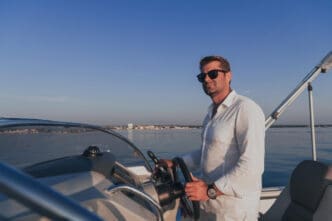 A focused senior executive, dressed in casual attire and sunglasses, relishes his vacation as he navigates a luxury boat during sunset