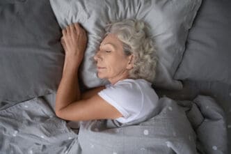 A serene, older woman with gray hair is peacefully sleeping on a soft pillow, tucked under a cozy blanket, viewed from above.