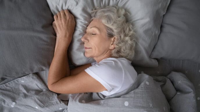 A serene, older woman with gray hair is peacefully sleeping on a soft pillow, tucked under a cozy blanket, viewed from above.